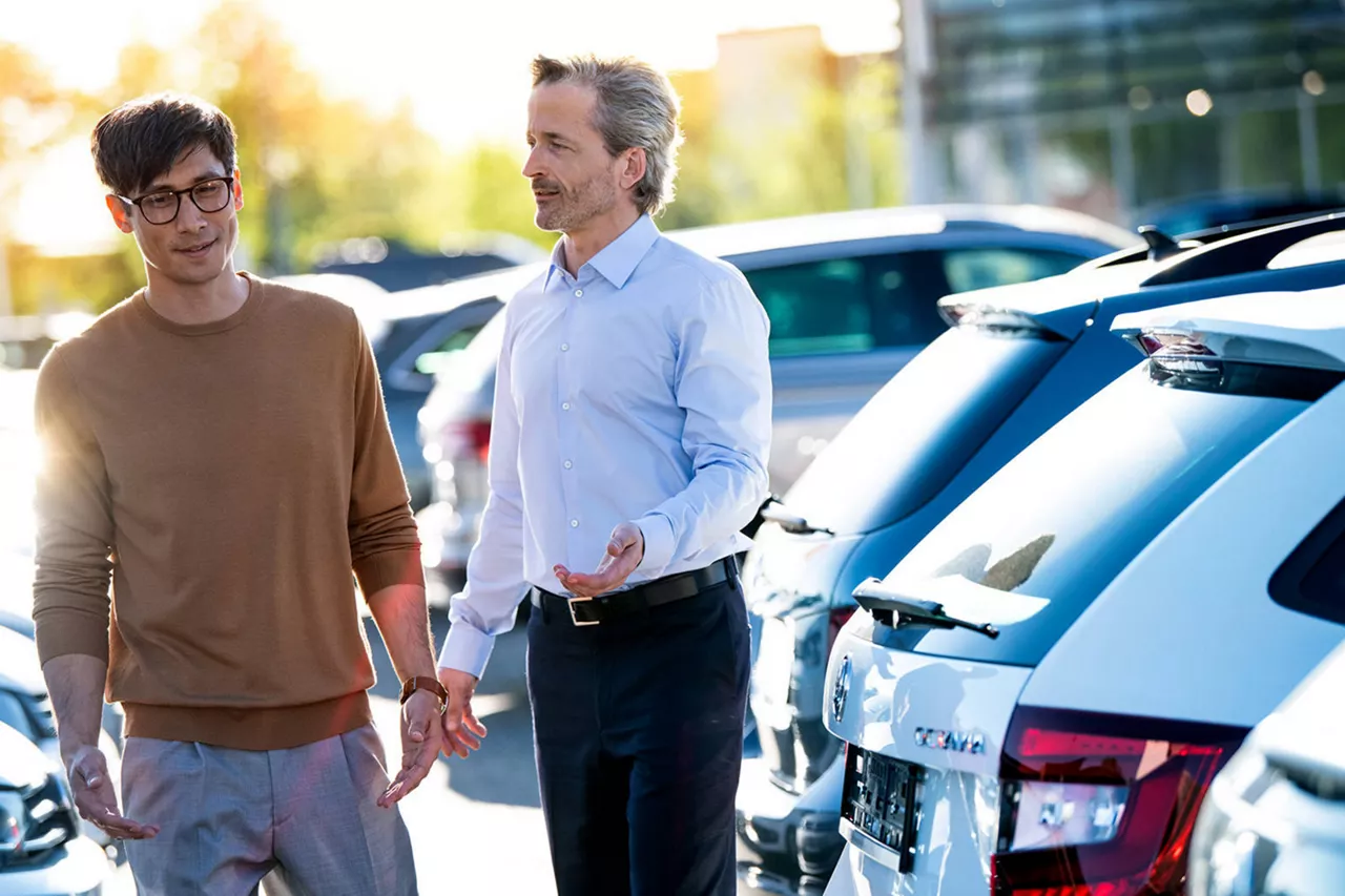 Dos hombres de pie hablando detrás de unos coches aparcados