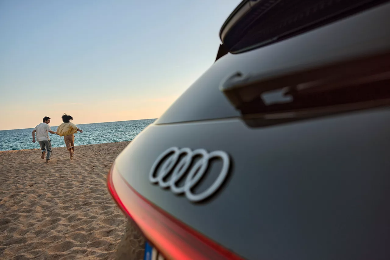 Zwei Personen laufen barfuß über einen Sandstrand Richtung Meer, im Vordergrund ein schwarzes Audi-Heck mit Logo.