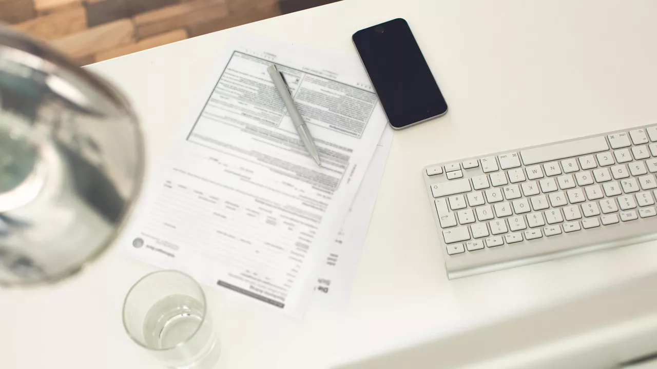 Auf einem Tisch liegen eine Bankvollmacht, ein Stift, ein Smartphone, eine Tastatur und ein Wasserglas.