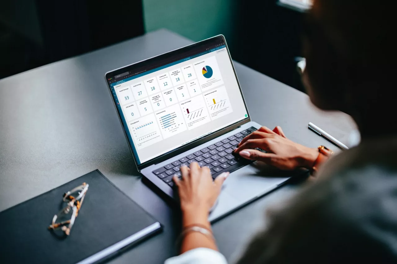Frau sitzt an einem Tisch und tippt auf einem geöffneten Laptop. Neben dem Laptop liegt ein zugeklapptes Heft und darauf eine Brille. Rechts daneben ein Stift.