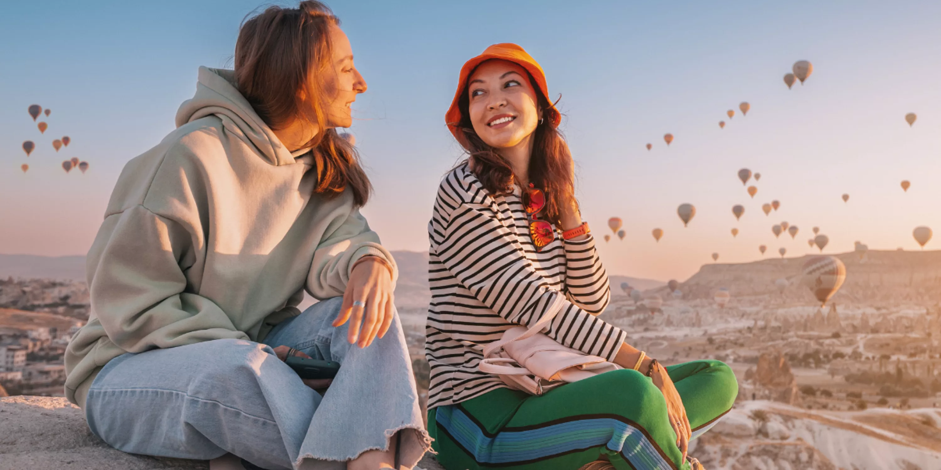 Zdjęcie pokazuje dwie uśmiechnięte kobiety na skale, a w tle widać balony startujące w Kapadocji.