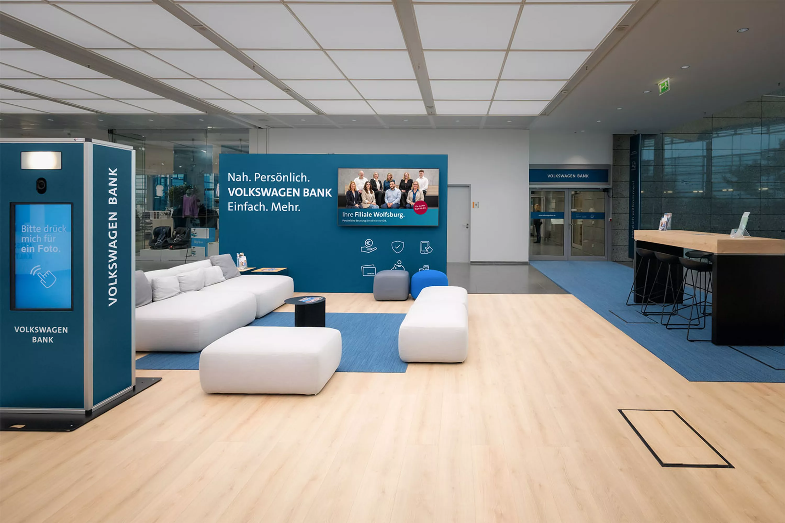 Moderne Empfangs- und Loungefläche der Volkswagen Bank mit Sitzgelegenheiten, Fotobox und Infowand. An der Wand hängt ein Bildschirm mit dem Team der Filiale Wolfsburg