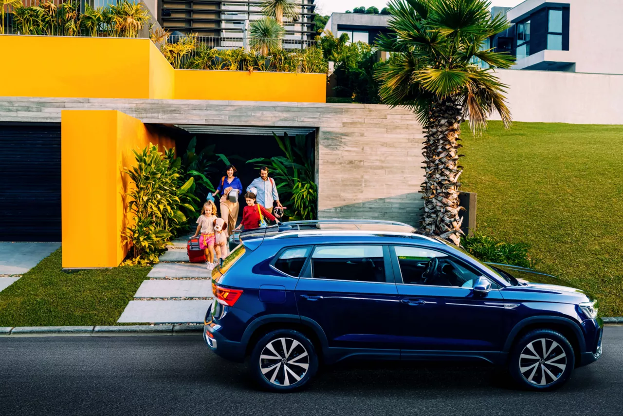 Carro estacionado em frente a uma casa. Ao fundo, vemos uma família caminhando em direção à ele. As crianças estão à frente, o casal está ao fundo. 