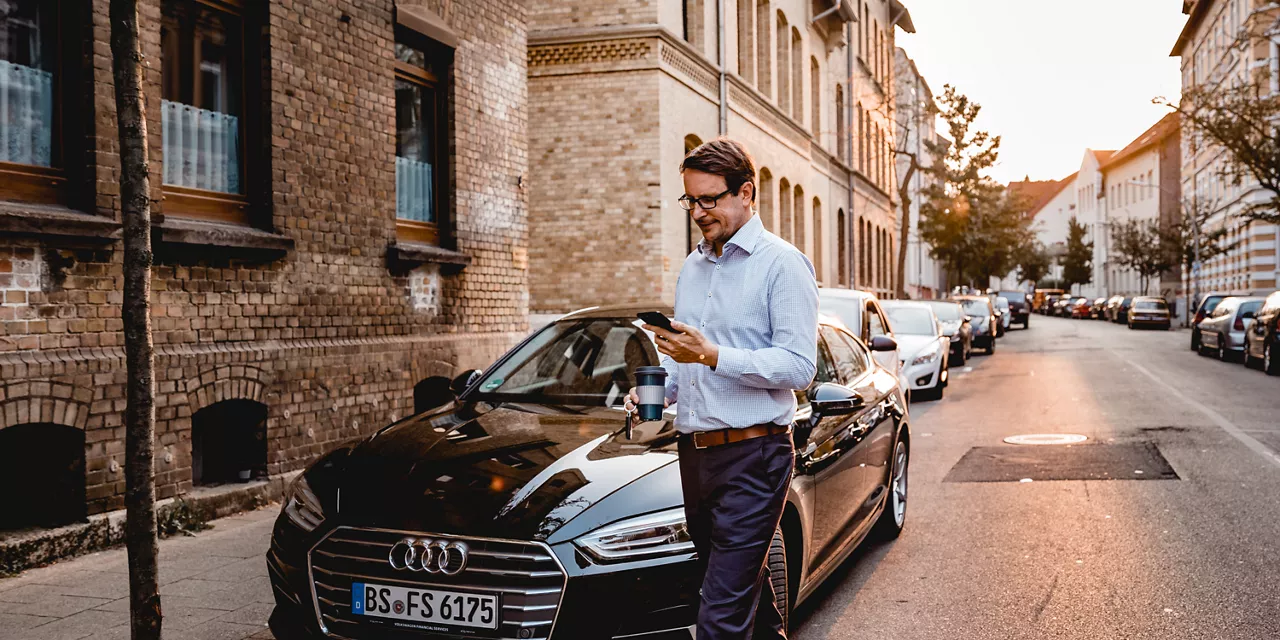 Homem caminhando em frente a um carro tomando café e olhando seu celular. 