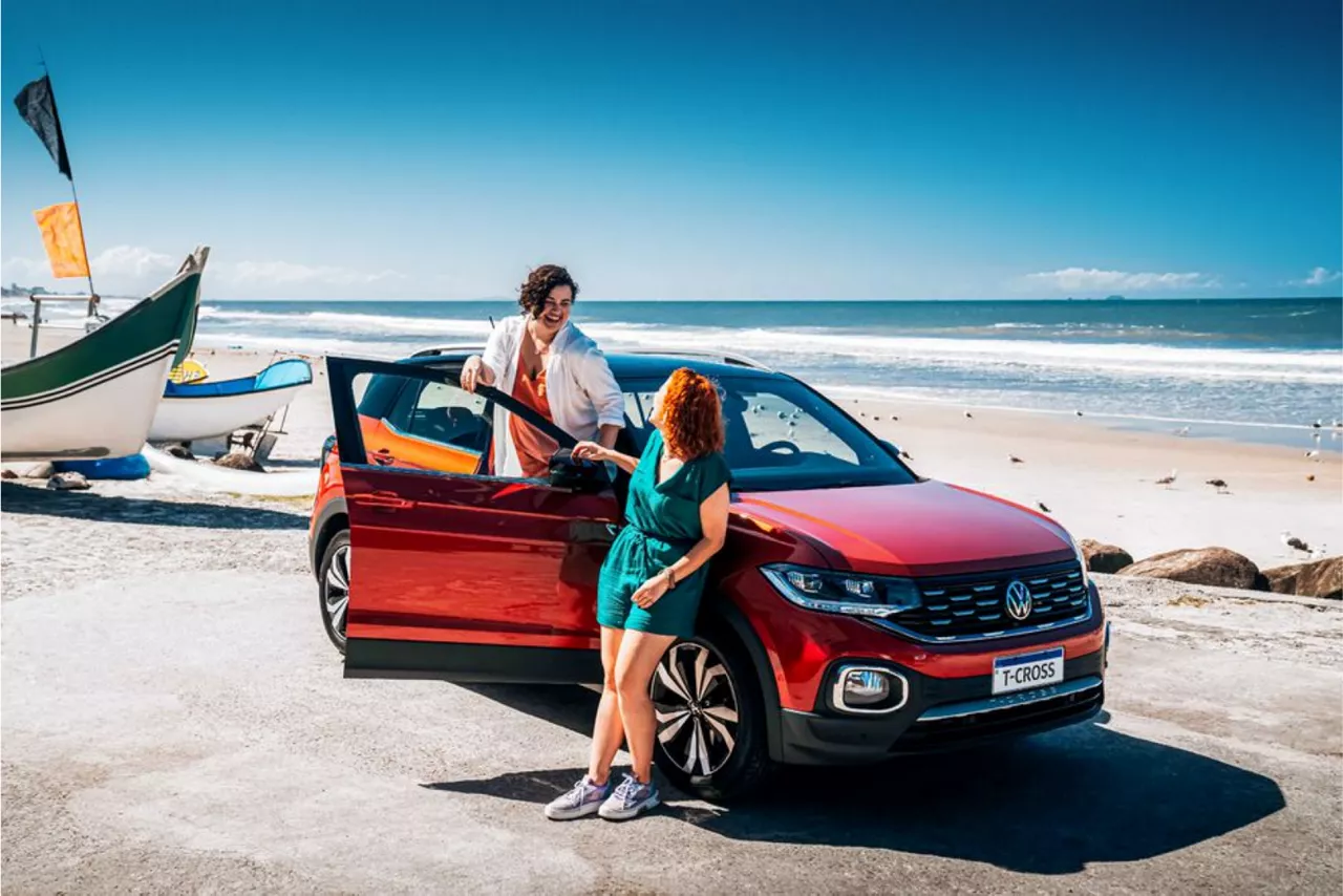 Carro estacionado em frente a uma praia. Tem uma mulher escorada na frente dele e a outra está em pé, com metade do corpo para dentro do carro e metade para fora, se segurando na porta do carro, que está aberta. 