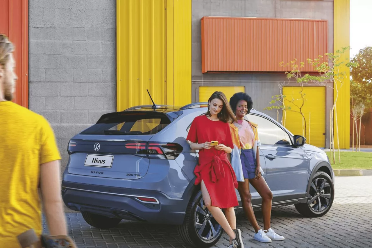 Duas mulheres escoradas em um carro estacionado na rua. Uma delas está olhando para o celular, a outra sorri enquanto um homem passa do outro lado da rua. 