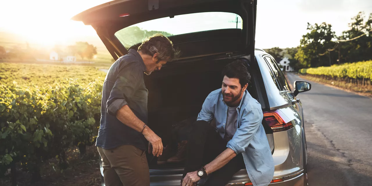 Dois homens em frente a um carro. Um deles está sentado no porta-malas, arrumando o tênis. 