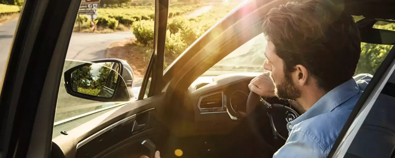 Homem dentro de um carro, no lugar do motorista, com uma mão no volante e a outra abrindo a porta. 