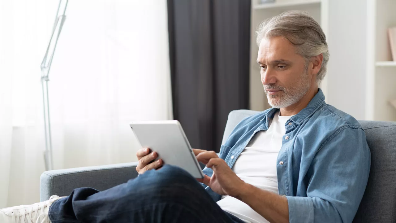 Mann mittleren Alters sitzt in einem hellen Wohnzimmer und hält ein Tablet in den Händen