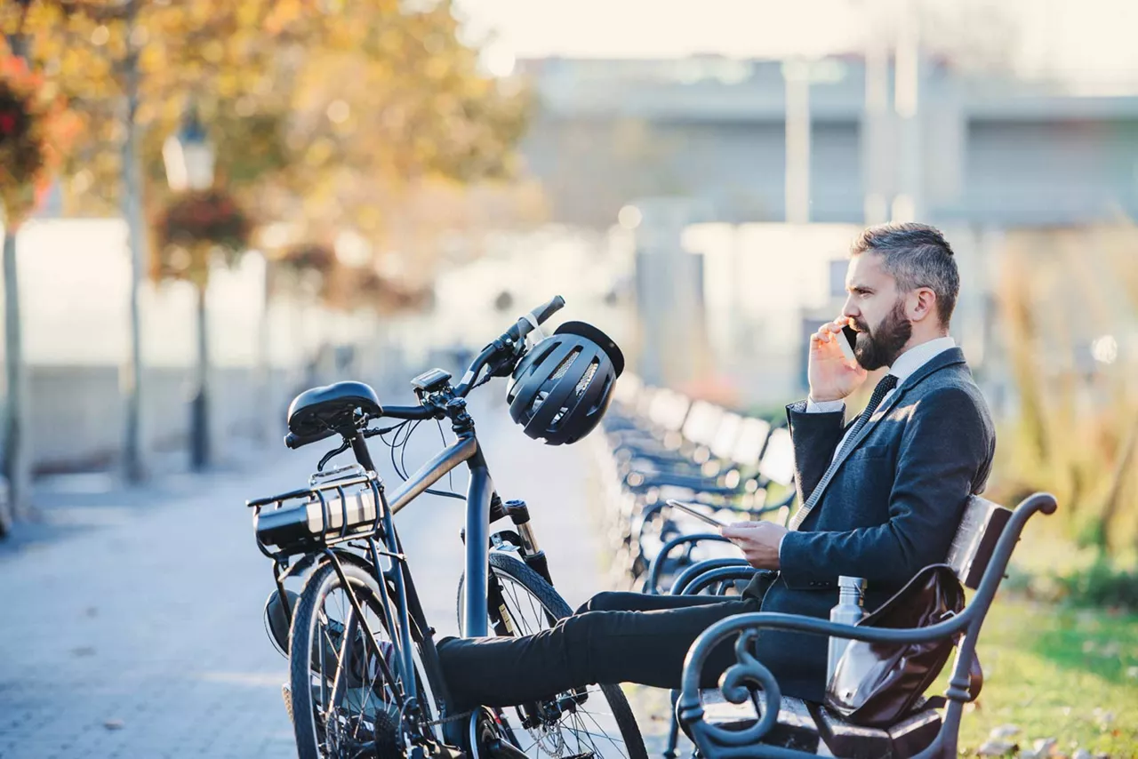 Geschäftsmann sitzt hinter seinem geleasten E-Bike auf einer Bank und telefoniert.