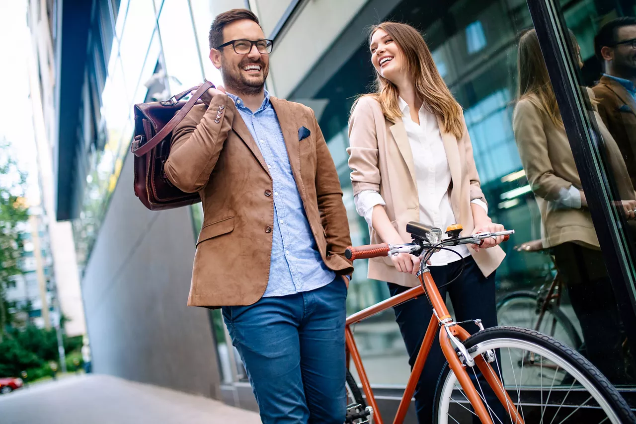 Ein Mann und eine Frau laufen lächelnd nebeneinander an einem großen Gebäude vorbei. Die Frau schiebt ein Fahrrad.
