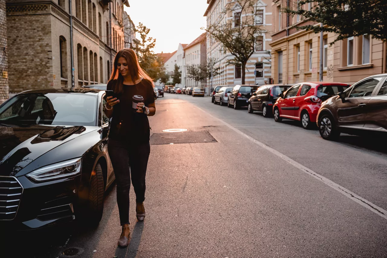 Mulher em pé com os braços abertos andando com um pé em frente ao outro. Ao fundo há um carro estacionado. 