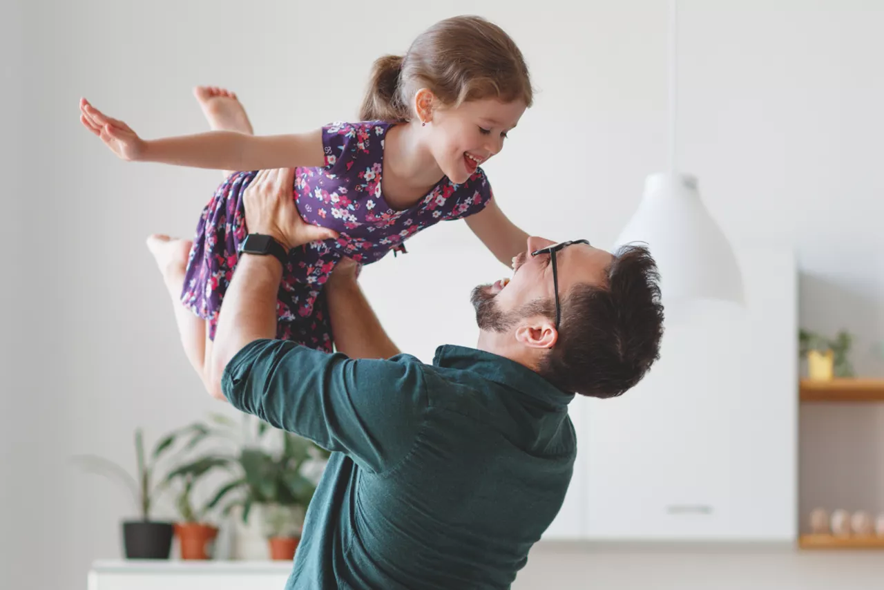 Pai e filha brincando. Ele está segurando ela para o alto. Ambos sorriem. 