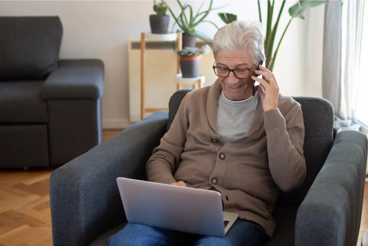 Homem sentado no sofá, falando no celular enquanto olha para um notebook. 