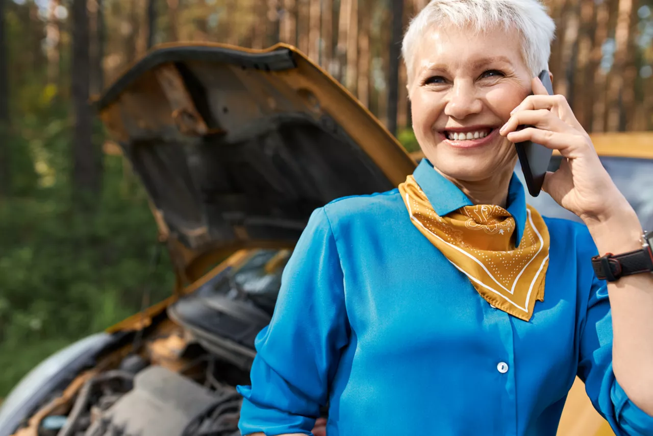 Seguro Volkswagen: mulher no primeiro plano da imagem sorrindo e falando ao celular. Ao fundo um carro com o capô aberto, mostrando que há algo de errado..