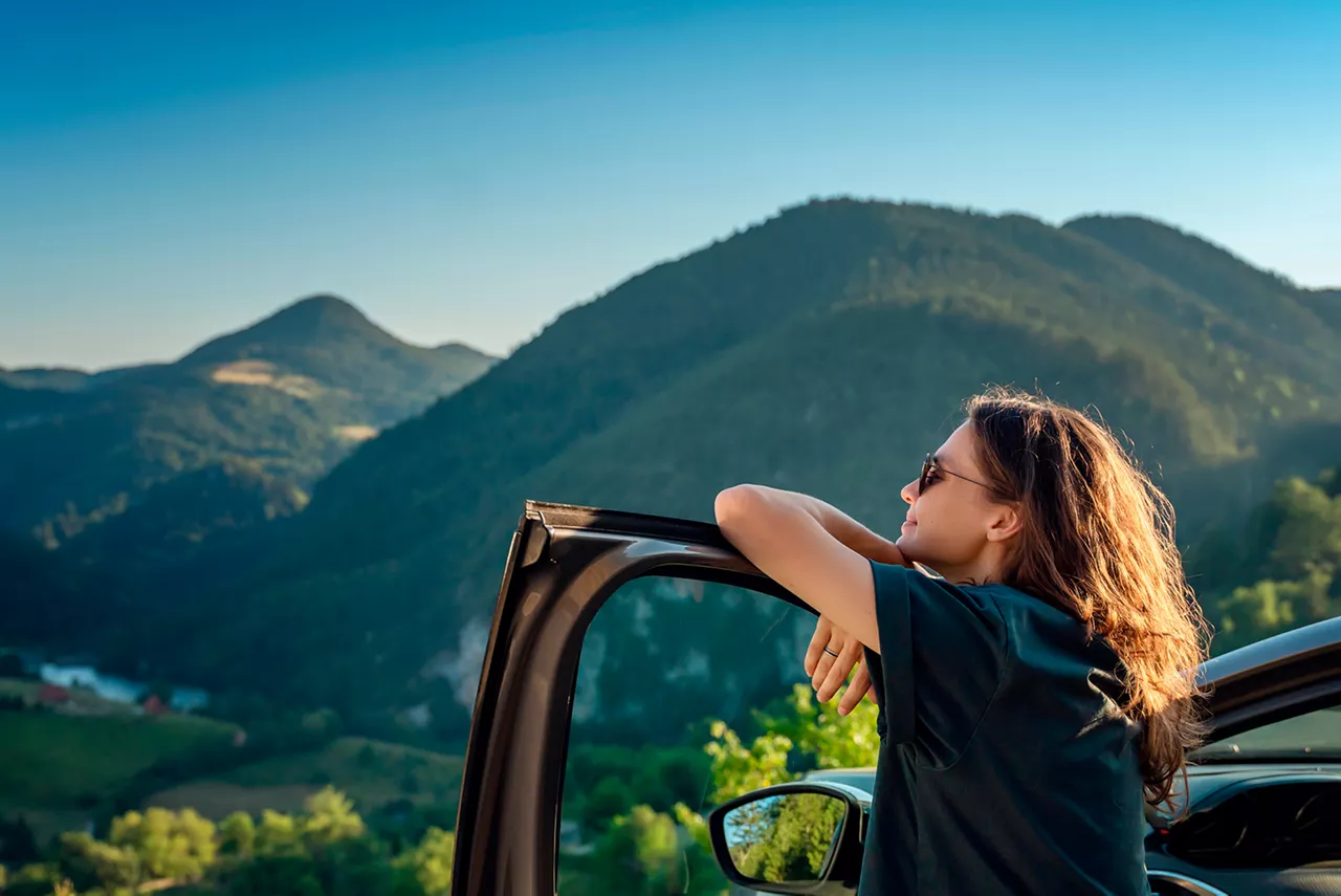 Una mujer apoyada en la puerta del conductor mirando unas montañas
