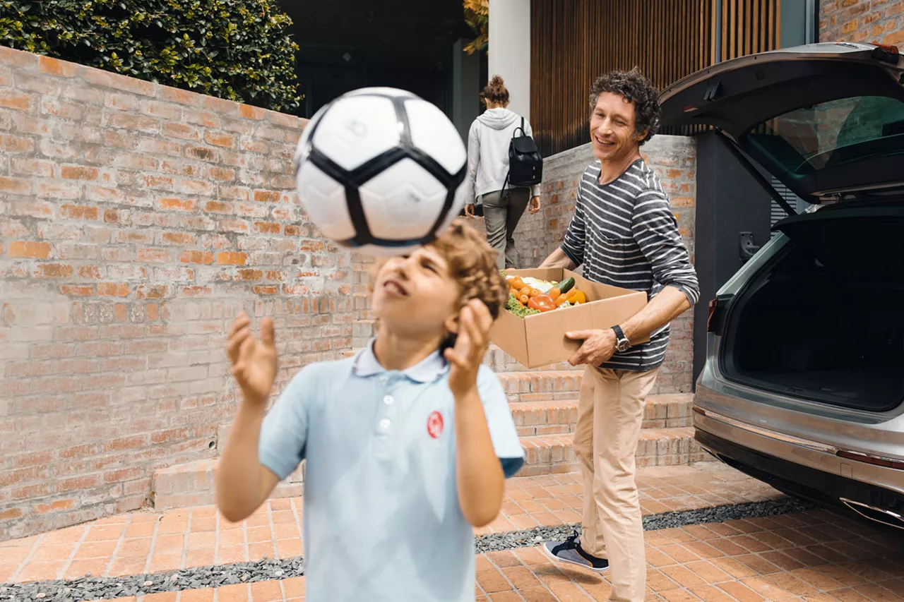 Plano medio de un niño jugando con un balón de fútbol, al fondo su padre saca del maletero del coche de alquiler del Grupo Volkswagen una caja con verduras.