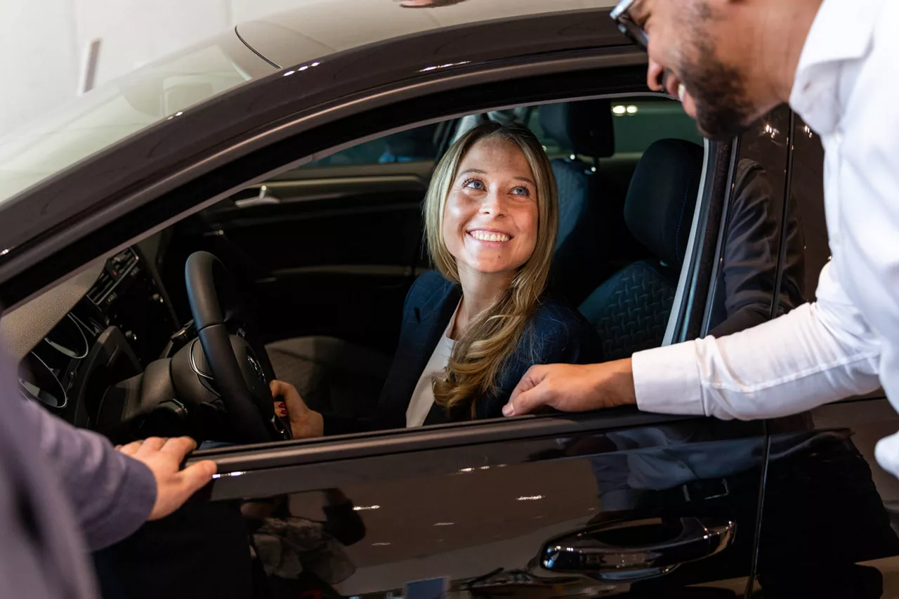 Una mujer sonriente sentada al volante de un coche de renting conversa con un hombre que está de pie junto a su coche. 