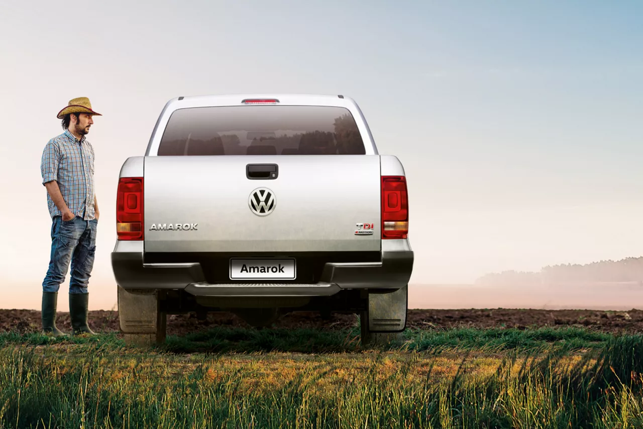 Foto de homem de pé ao lado de uma caminhonete Amarok. A caminhonete está de costas, e é prata. O homem está de lado olhando para ela. Ele usa chapéu de campo, camisa xadrez, calça jeans e botas. A paisagem é céu aberto e claro, o chão é de mato baixo. Fim da descrição.