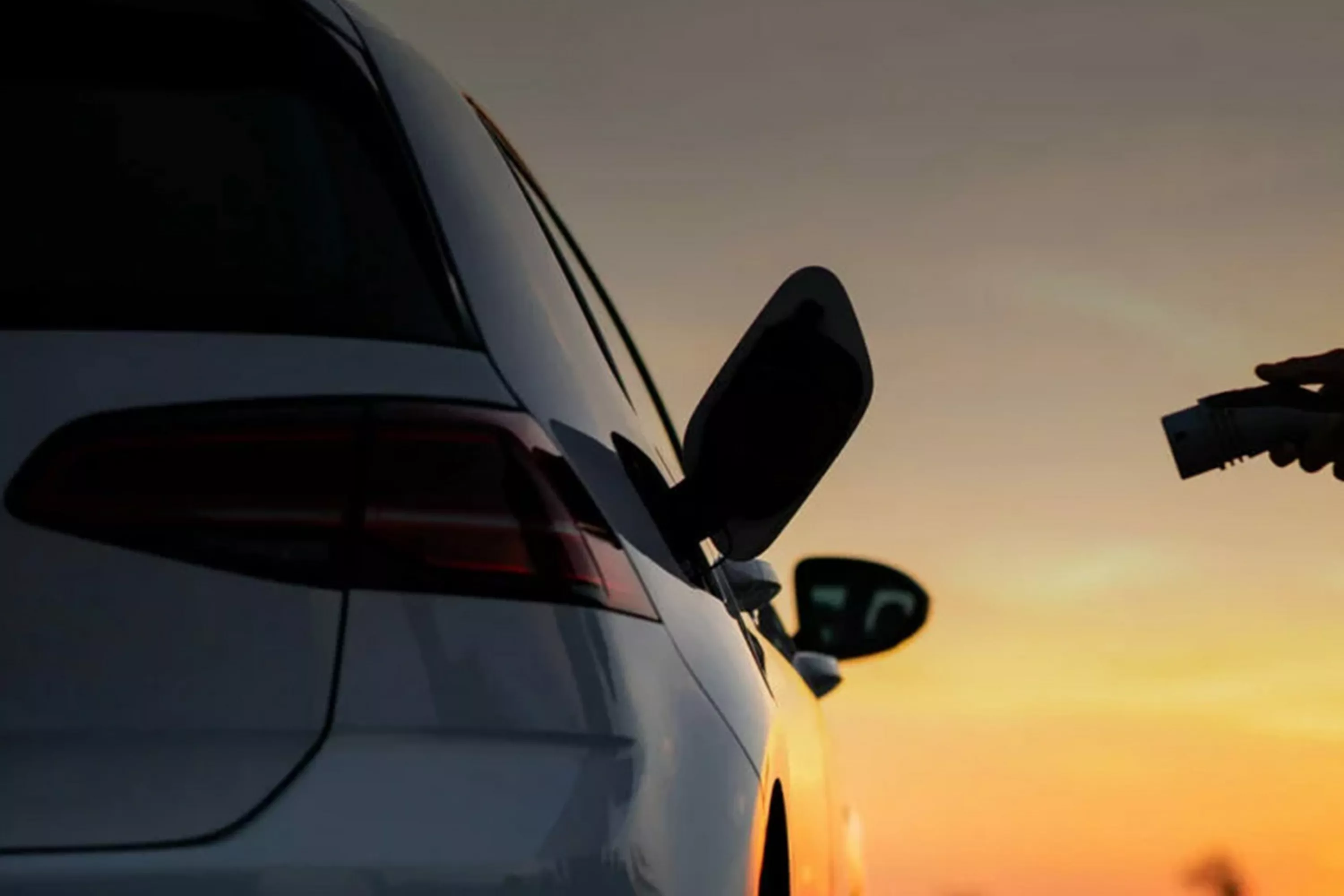 Eine Person hält einen Stecker neben einem E-Fahrzeug. Im Hintergrund ist ein Sonnenuntergang zu sehen.