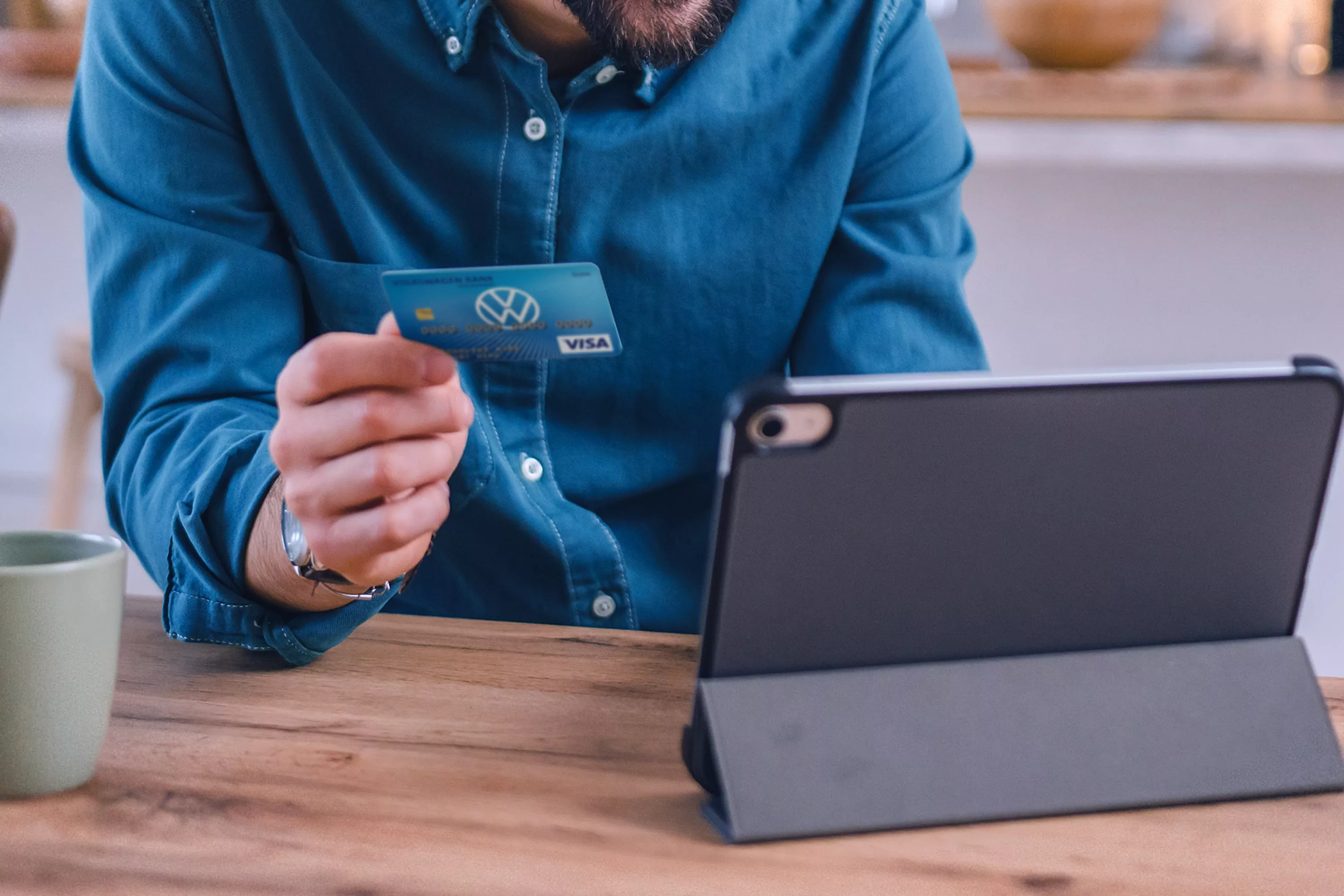 Mężczyzna trzymający kartę bankową w jednej dłoni, na stole leży urządzenie typu tablet.