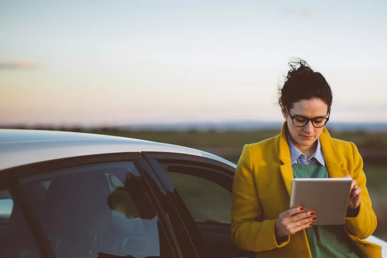 Una mujer con una tablet apoyada en un coche al atardecer