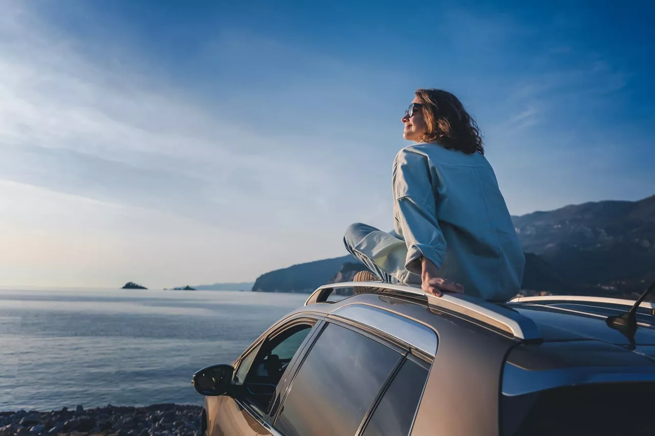 Una chica sentada en el techo de un coche de alquiler mirando el mar, VW FS | Rent-a-Car, en la playa