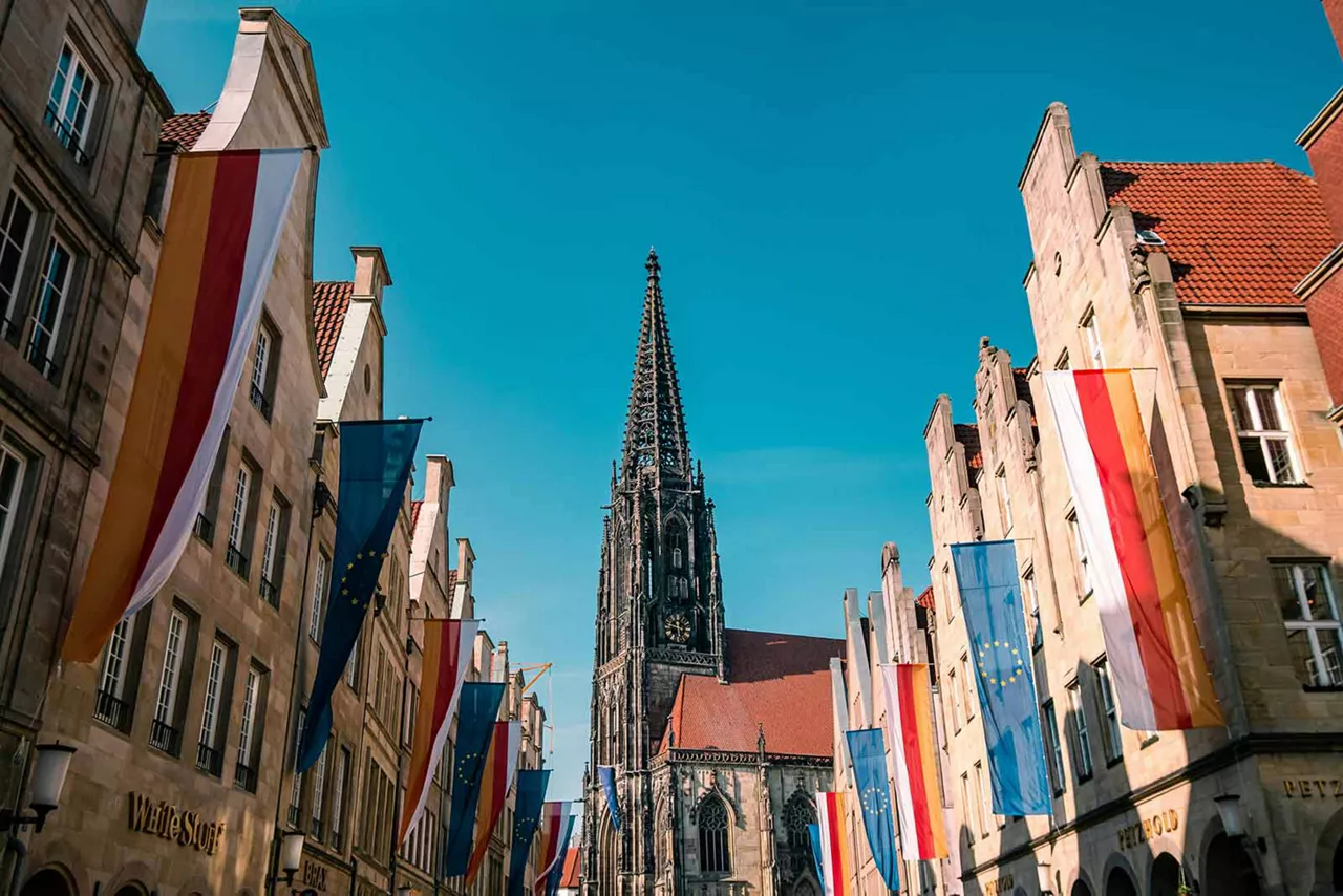 Historische Straße mit Fahnen und Blick auf den Kirchturm des St.-Paulus-Doms in Münster bei blauem Himmel