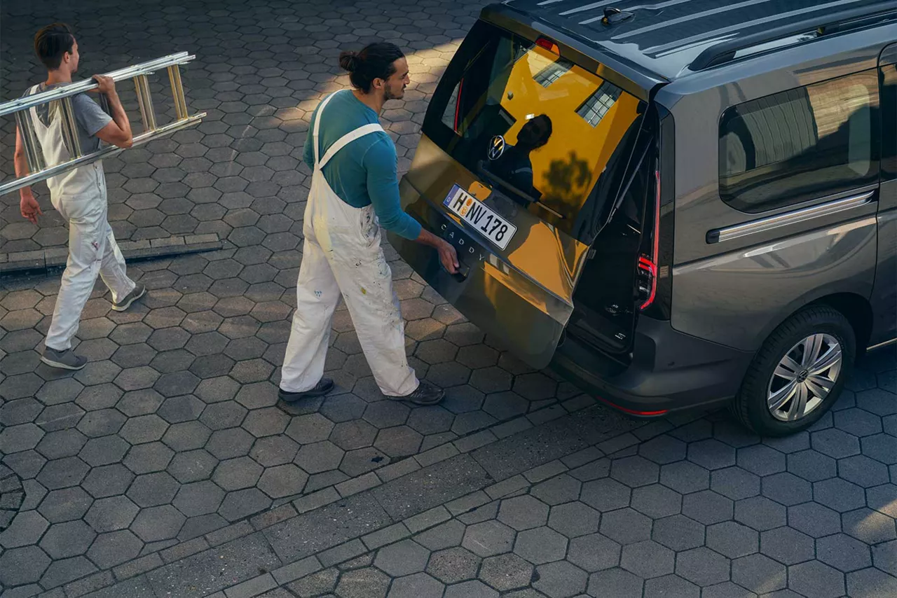 handwerker-in-latzhose-öffnet-kofferraum-eines-grauen-vw-caddy