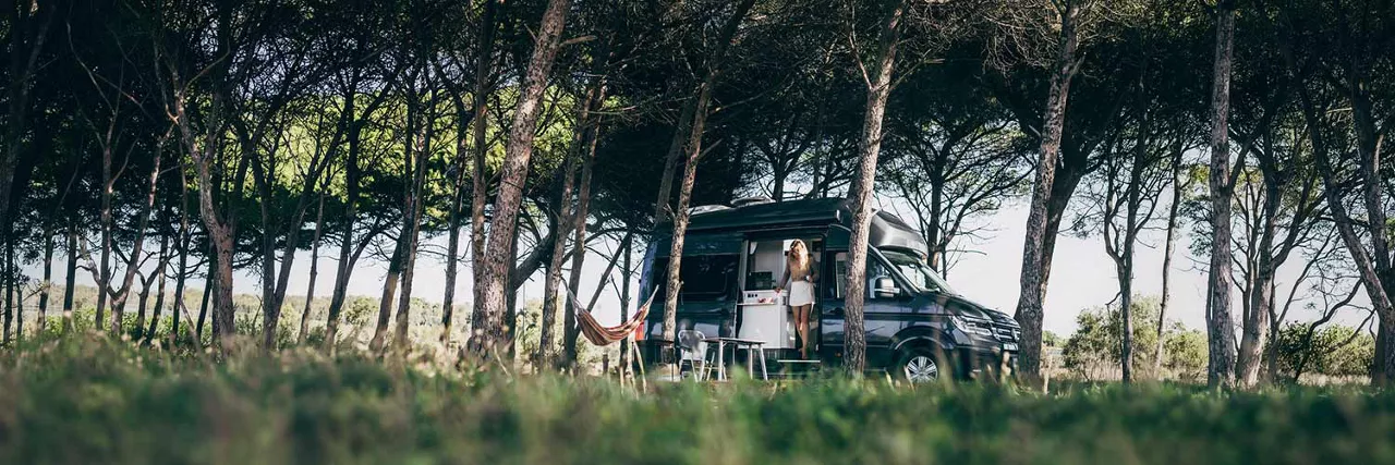 VW Grand California mit geöffneter Schiebetür steht in naturnaher Waldumgebung, im Vordergrund eine Hängematte und Campingausstattung