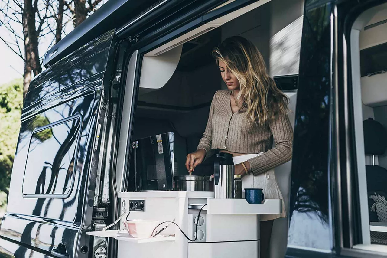 Person bereitet Kaffee in der geöffneten Küche eines VW Campervans in naturnaher Umgebung zu.