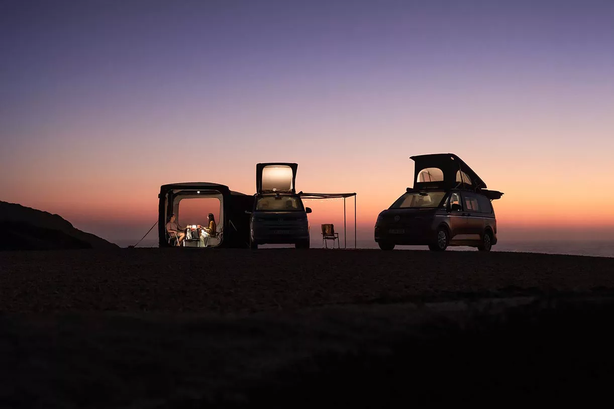  Zwei Camper stehen bei Sonnenuntergang; im Vorzelt sitzen zwei Personen, innen ist Licht an.
