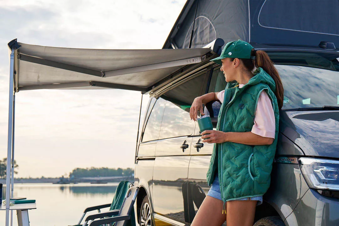 Person lehnt an einem Camper unter einer ausgefahrenen Markise und hält einen Becher, mit Blick auf einen See im Hintergrund