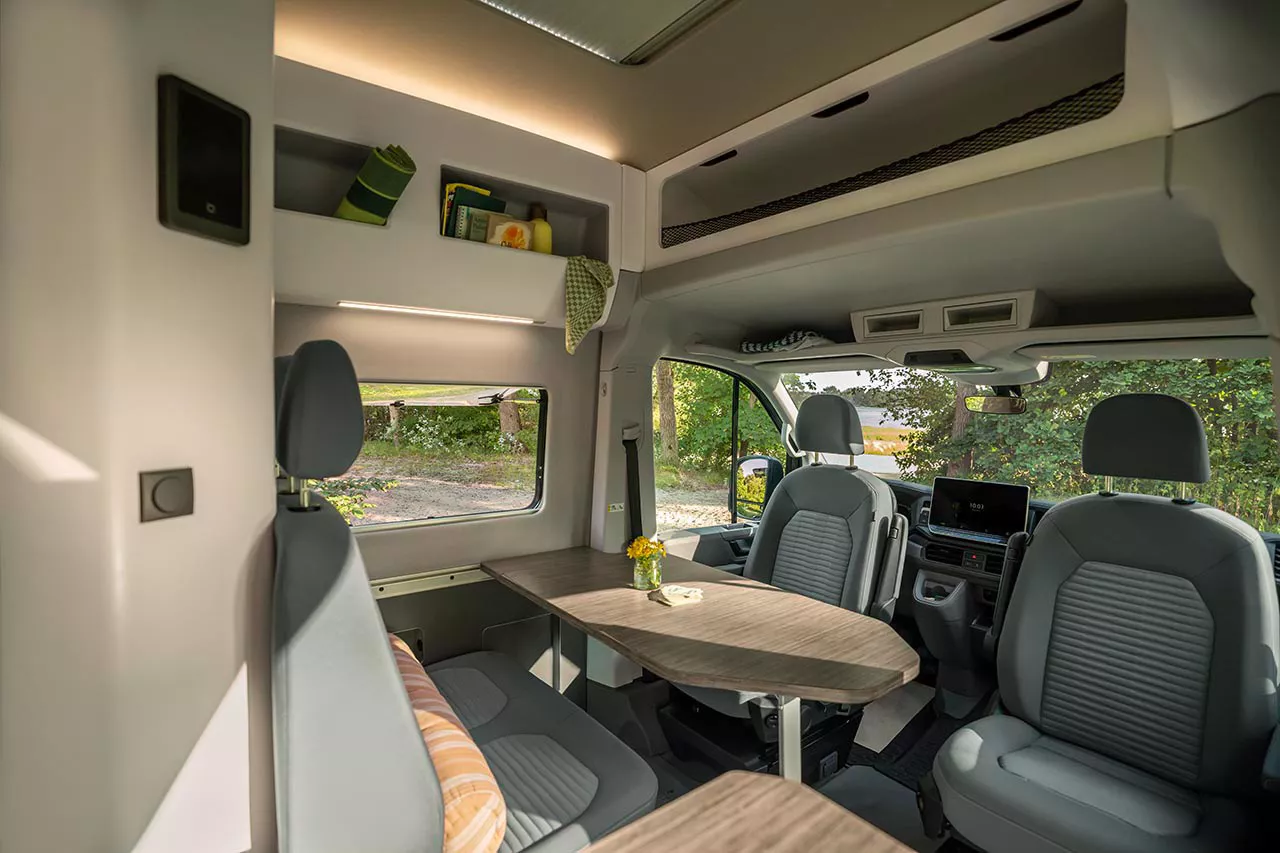 Innenraum eines Campers mit Sitzgruppe, drehbaren Vordersitzen und einem klappbaren Tisch; Tageslicht fällt durch Fenster auf einen Stellplatz mit Bäumen.