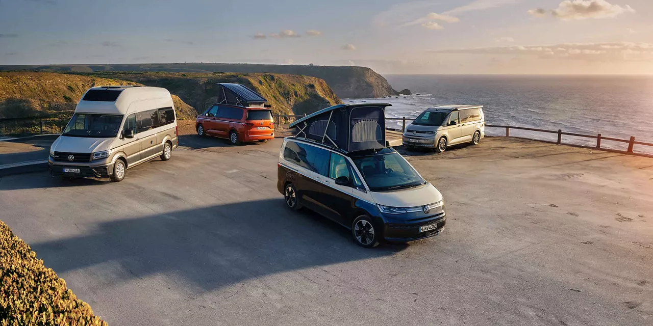 Mehrere VW-Campervans mit geöffneten Aufstelldächern stehen auf einem Parkplatz an einer steilen Küstenlandschaft mit Blick auf das Meer bei Sonnenuntergang