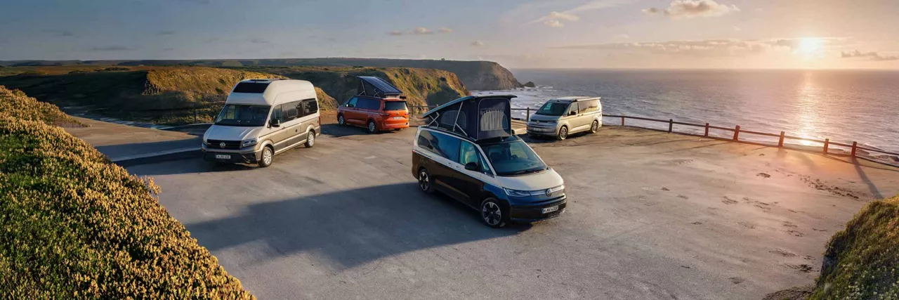 Mehrere VW-Campervans mit geöffneten Aufstelldächern stehen auf einem Parkplatz an einer steilen Küstenlandschaft mit Blick auf das Meer bei Sonnenuntergang