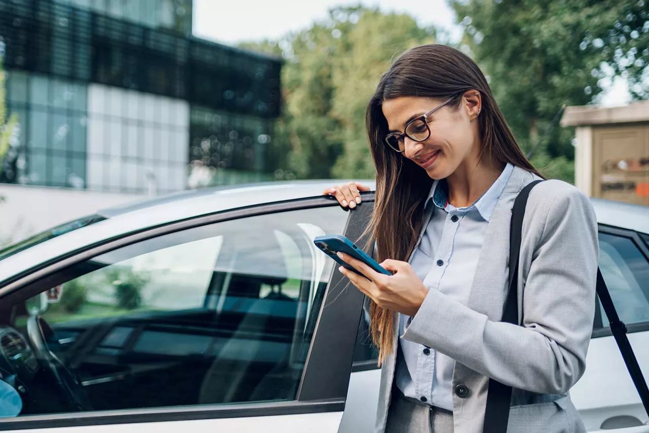 Lächelnde Frau öffnet Autotür und schaut auf ihr Smartphone