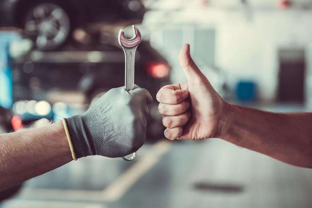 Hand mit Schraubenschlüssel und Hand mit Daumen hoch nebeneinander in einer Autowerkstatt