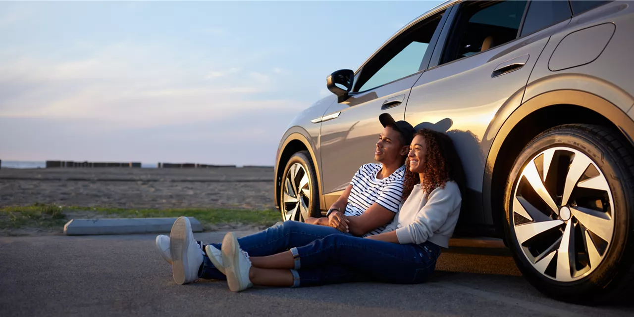 Na foto, há um carro cinza estacionado próximo a um campo aberto com céu azul ao fundo. Há dois jovens, uma mulher e um homem, que sorriem sentados no chão e encostados na porta traseira esquerda do veículo, olhando para o horizonte e recebendo uma luz dourada na face. A mulher é negra, tem cabelos cacheados e avermelhados na altura dos ombros, veste blusa cinza de mangas compridas, calça jeans azul e tênis branco. O homem é negro e sem barba, usa boné preto, camiseta com listras brancas e azuis, calça jeans azul e tênis branco. Na lateral superior esquerda encontra-se a marca corporativa da Volkswagen Financial Services escrito em letras maiúsculas brancas largas dentro de um quadrado azul degradê. Abaixo do título da marca está escrito em letras maiúsculas, menores e finas o slogan da companhia The key do mobility. À esquerda da foto no canto superior abaixo da marca corporativa está o título Relatório Anual. Menu de navegação na parte superior. Fim da audiodescrição