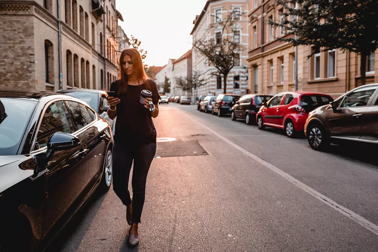 Frau schaut auf ihr Smartphone und läuft an einem Auto vorbei. Im Hintergrund sind eine Straße mit parkenden Autos und Gebäude am Straßenrand zu sehen.
