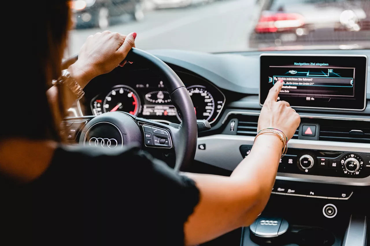Nahaufnahme vom Fahrersitz eines Autos von hinten: Eine Frau tippt auf das Navigationssystem auf dem Bildschirm in der Konsole des Autos. Mit der linken Hand hält sie das Lenkrad.