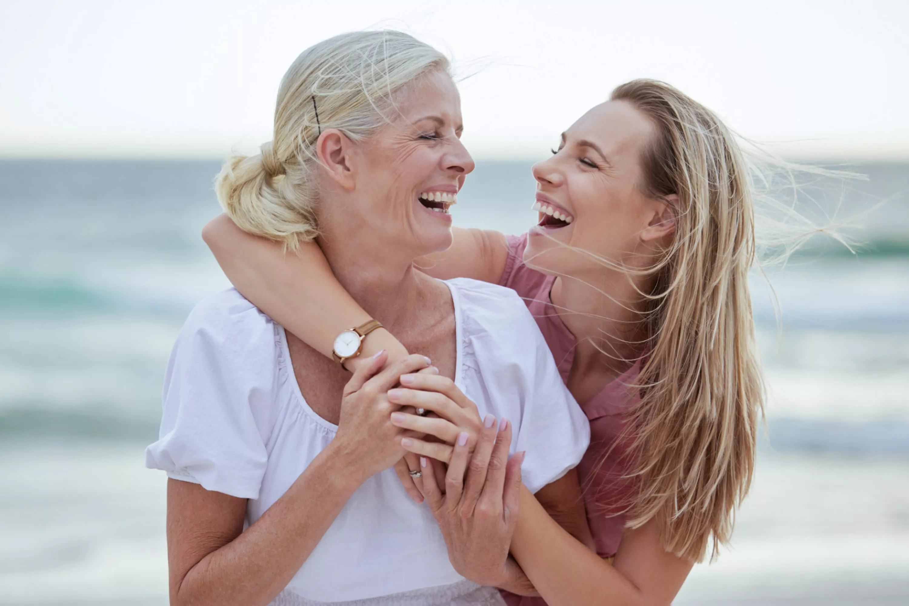 Frau umarmt eine ältere Frau von hinten. Beide schauen sich an und lachen. Im Hintergrund ist ein Strand zu sehen.