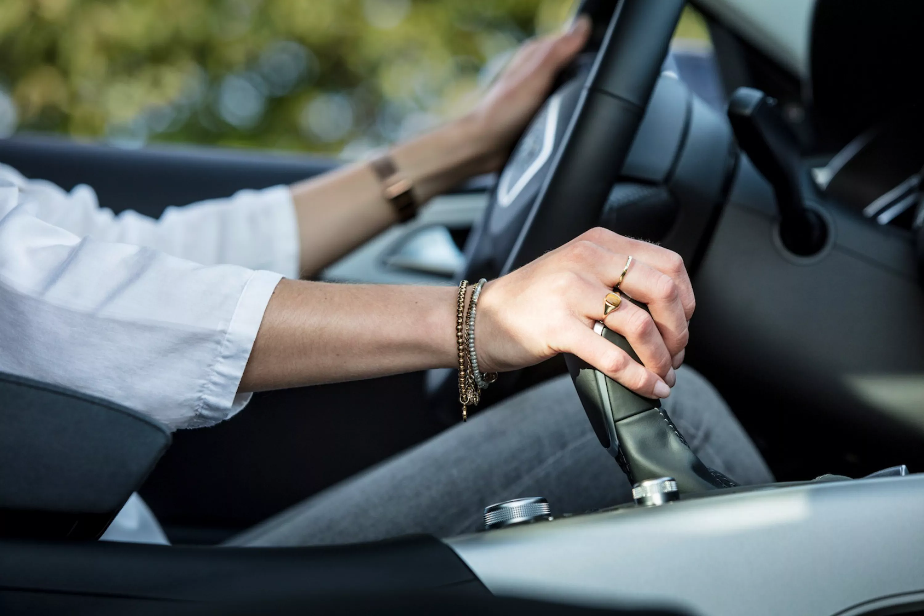 Nahaufnahme seitlich vom Fahrersitz in einem Auto. Eine Frau fährt und hält eine Hand am Lenkrad, die andere an der Schaltung.