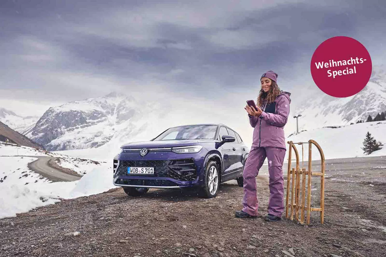 Frau in Winterkleidung steht neben einem VW Tayron in verschneiter Berglandschaft und hält ein Smartphone in der Hand. Daneben steht ein Holzschlitten.