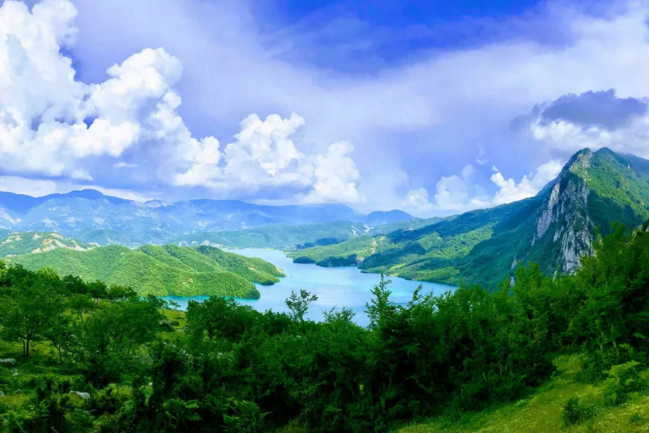  Grüne Berglandschaft mit See, bewaldeten Hügeln und blauem Himmel mit weißen Wolken in Albanien