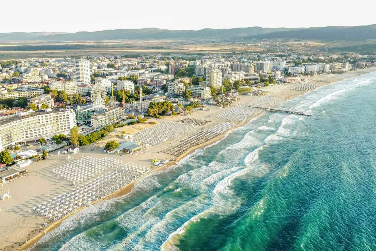 Luftaufnahme einer Küstenstadt in Bulgarien mit langem Sandstrand, geordneten Strandliegen, Meereswellen und Stadtbebauung entlang der Promenade