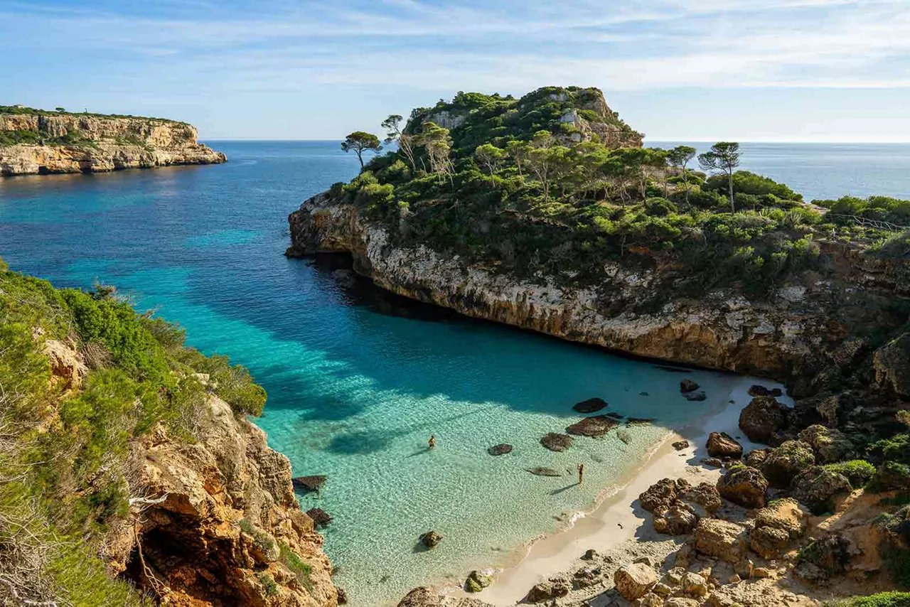 Türkisfarbene Badebucht mit Sandstrand, Felsen, klaren Wasser und bewachsenen Klippen an der Küste von Palma de Mallorca