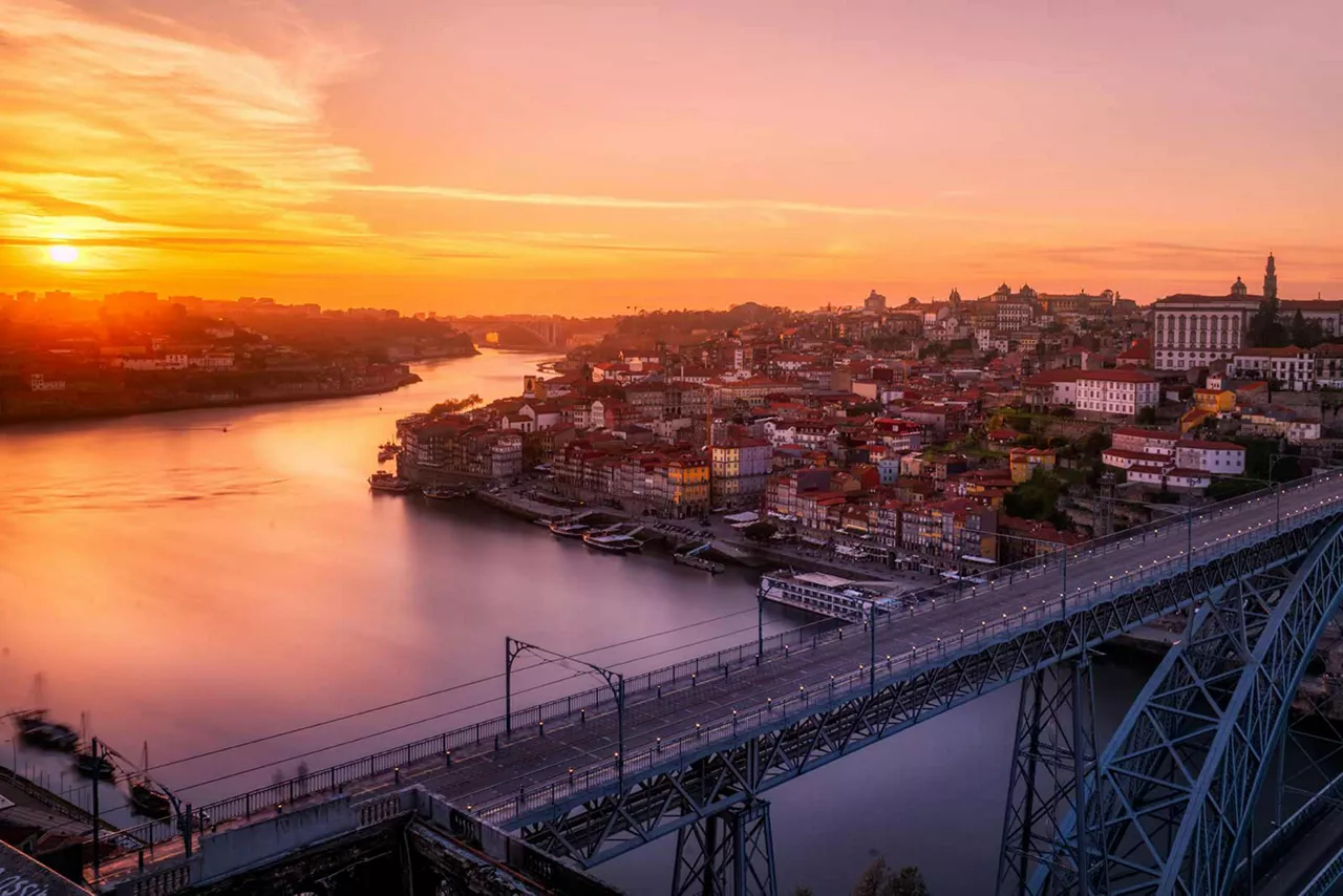 Stadtansicht über Porto mit Fluss und Brücke bei Sonnenuntergang, dicht bebautes Ufer und historische Gebäude im warmen Abendlicht