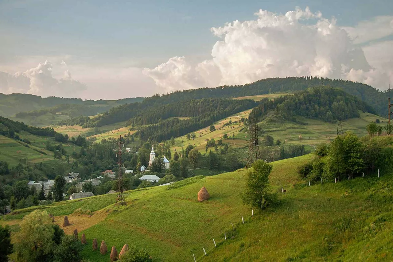 Hügelige Landschaft mit grünen Wiesen, bewaldeten Hängen, verstreuten Häusern und einer Kirche im Tal in Rumänien