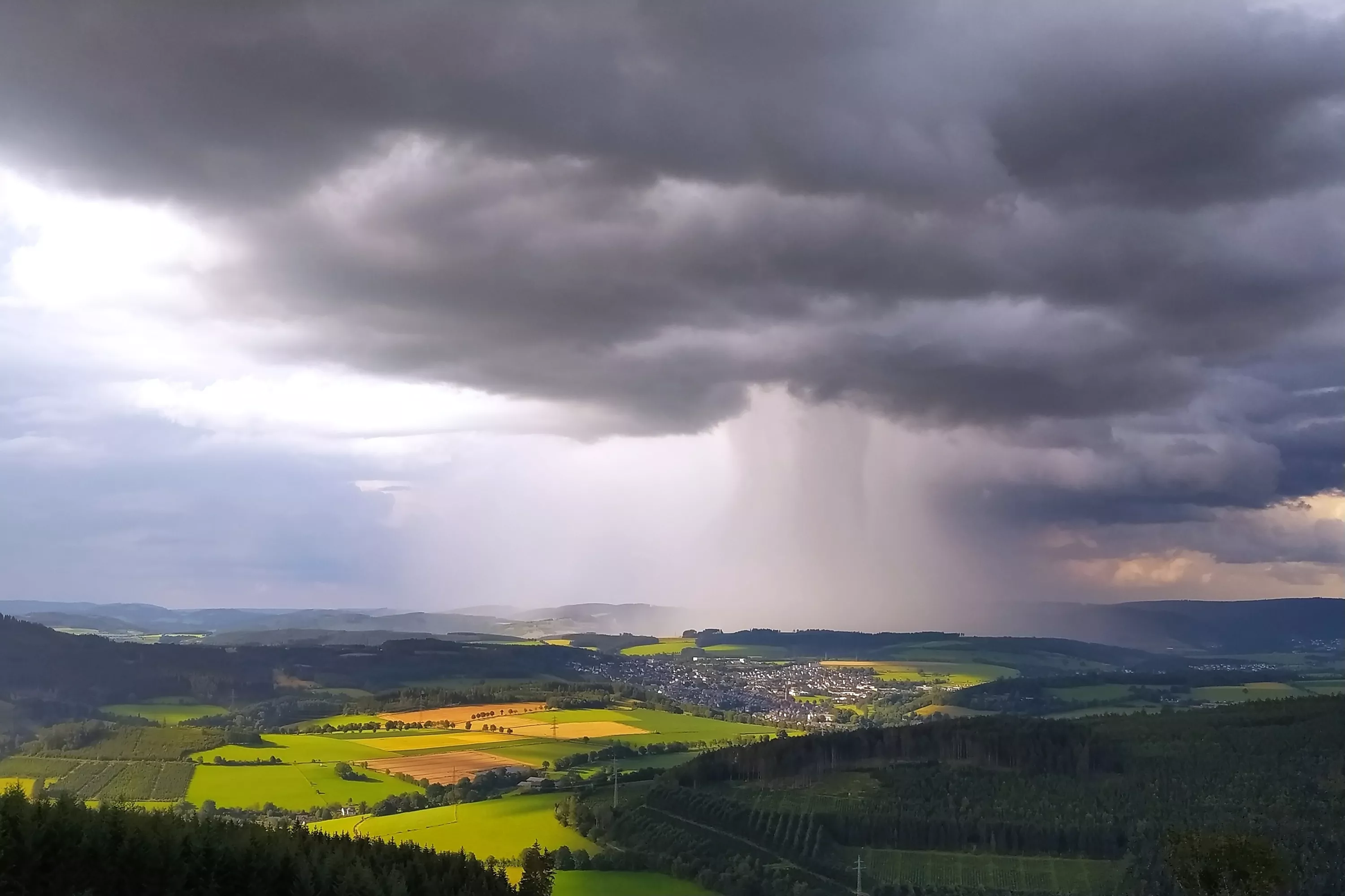 Dunkle Gewitterwolken mit starkem Regenschauer über einer ländlichen Landschaft mit Feldern und kleinem Ort.